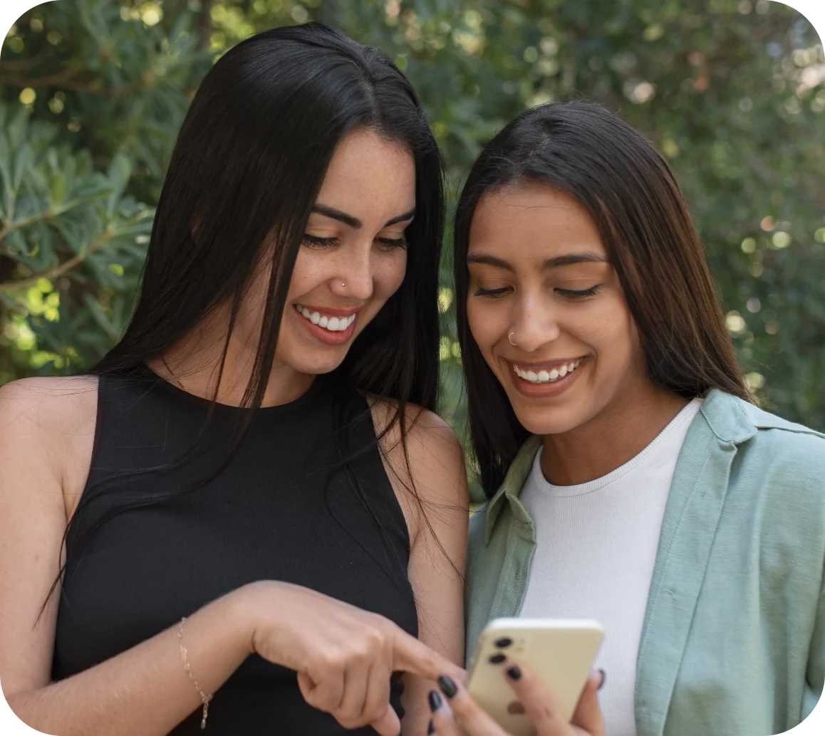 Two women stand closely together outdoors, both smiling and looking at a smartphone that one of them is holding.